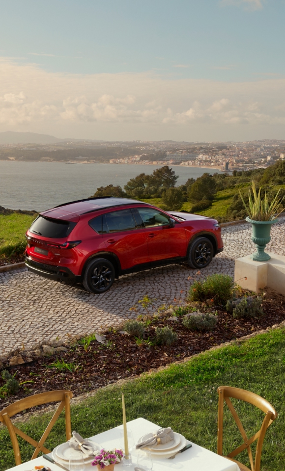 2026 Mazda CX-5 in Soul Red Crystal parked on a European cobblestone driveway with scenic sea view, black alloy wheels, and outdoor dining setup—luxury SUV lifestyle in coastal countryside setting.