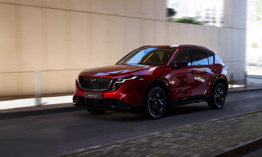 2026 Mazda CX-5 in Soul Red Crystal driving through urban streets, showcasing signature front grille with Mazda logo and CX-5 license plate—dynamic SUV performance in city environment.